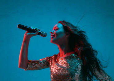 Caucasian female singer portrait isolated on blue studio background in neon light. Beautiful female model in bright dress with microphone. Concept of human emotions, facial expression, ad, music, art.